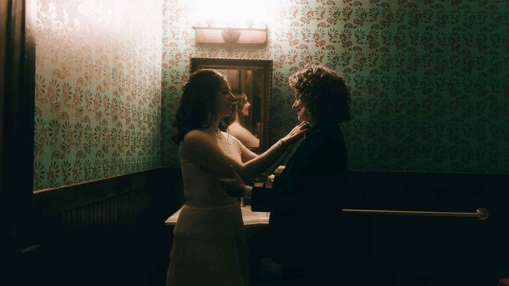 Two brides face to face in a papered washroom, one reaching up to straighten the other's collar