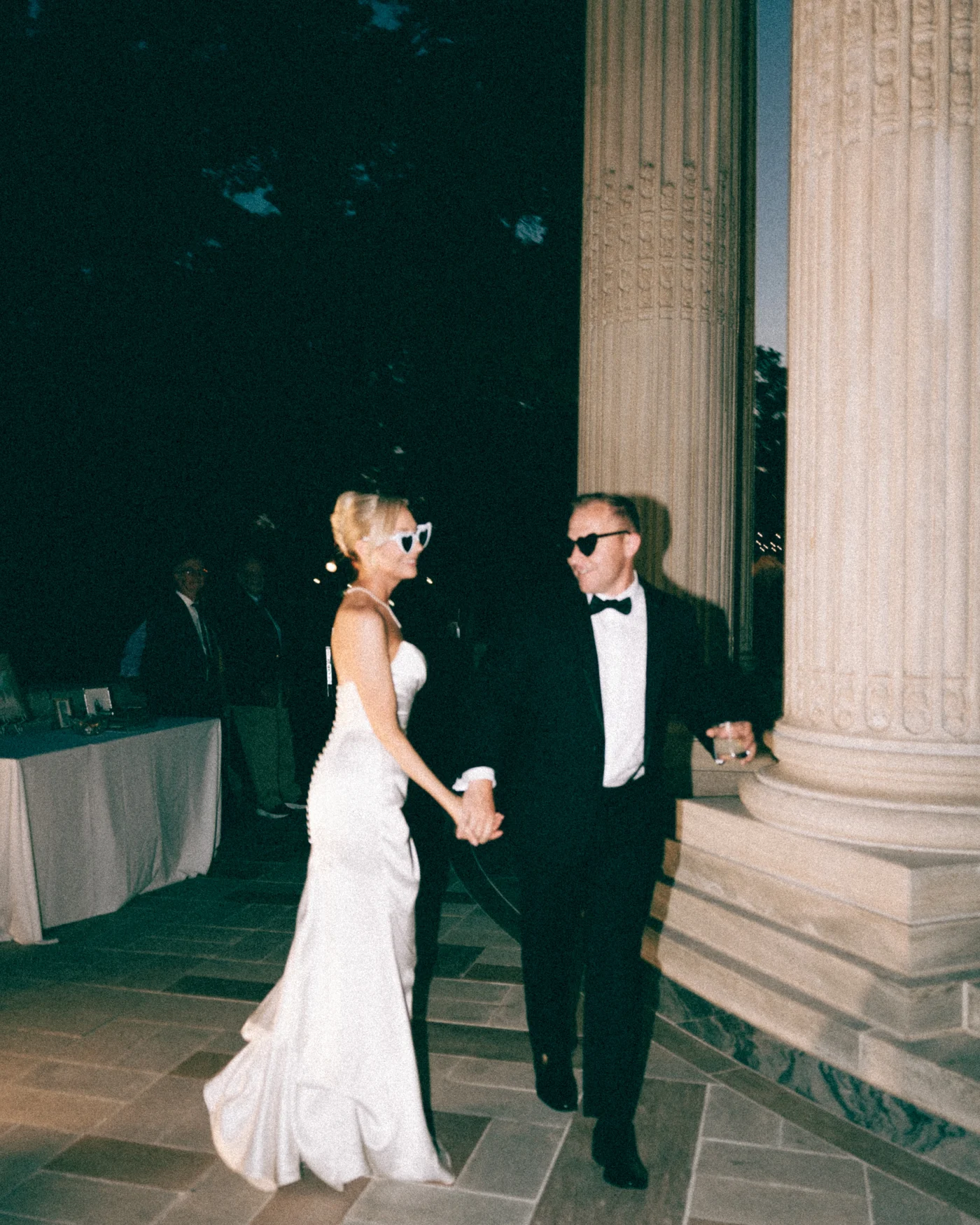 A couple in sunglasses walking a flagstone terrace at dusk, their guests trailing behind them between the columns