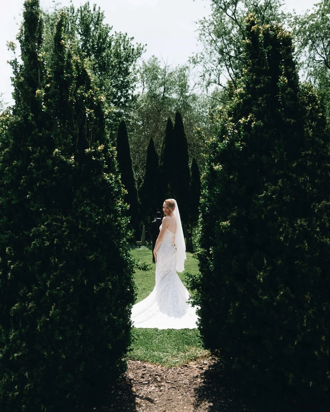 A bride standing alone at the end of a corridor of tall clipped hedges