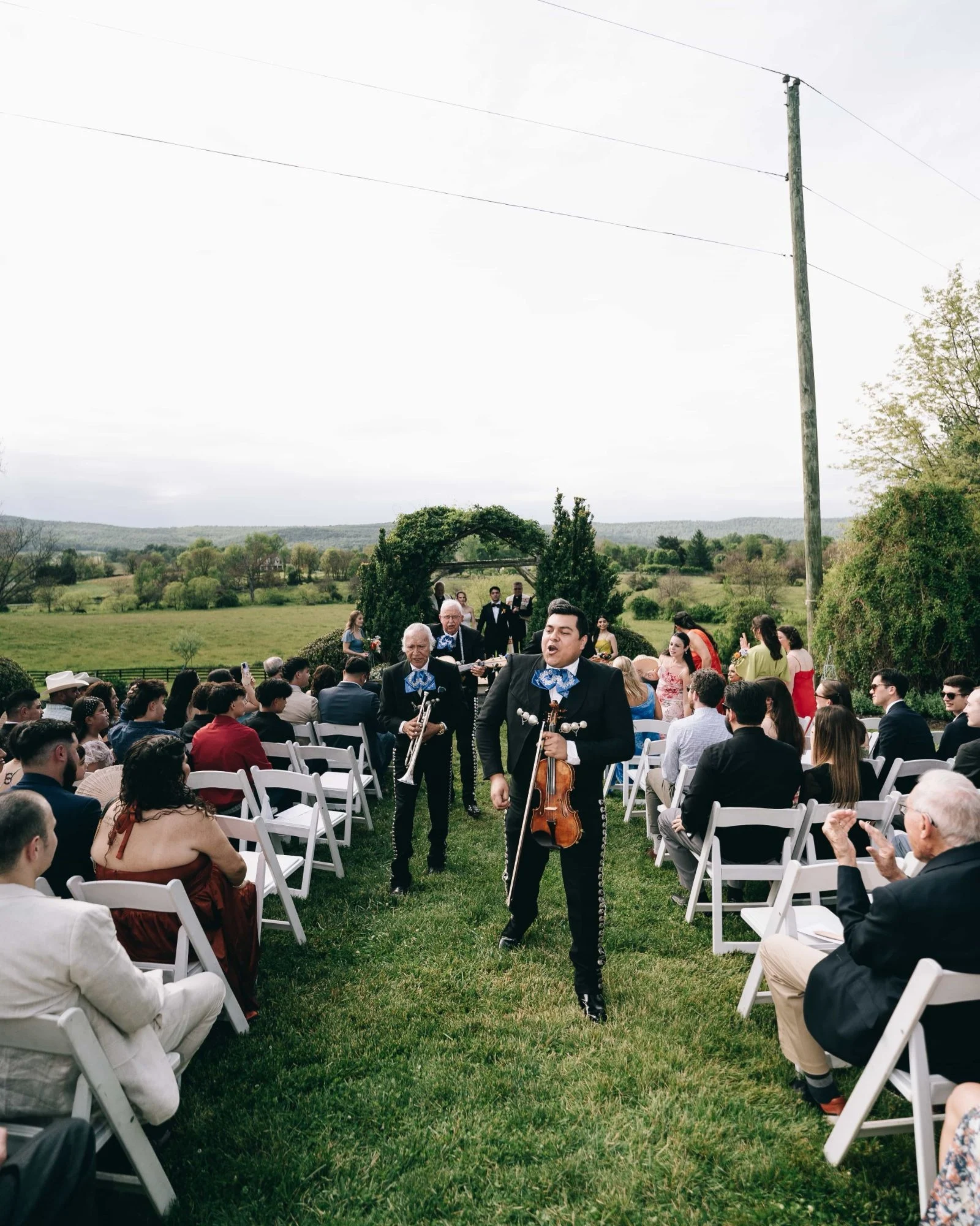 Mariachi musicians singing their way down the grass aisle of a hilltop ceremony, guests turned in their chairs to watch