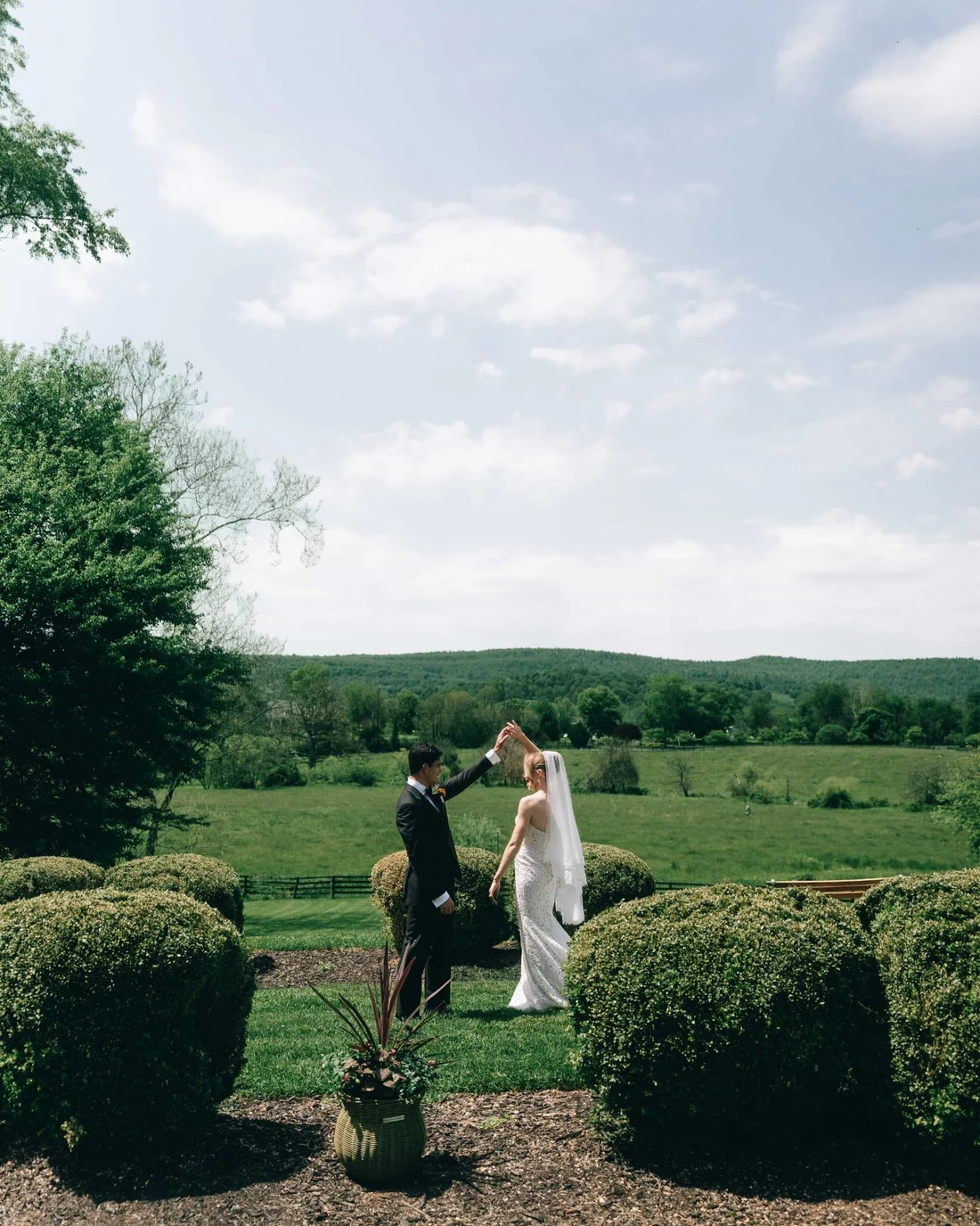 A couple alone on a lawn between clipped boxwoods, a wide green valley and a bigger sky behind them