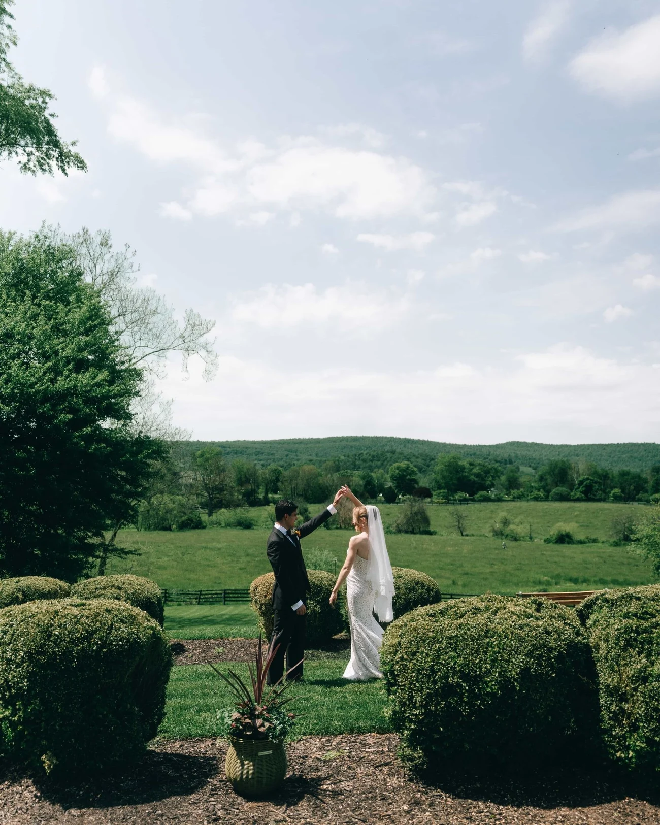 A couple alone on a lawn between clipped boxwoods, a wide green valley and a bigger sky behind them