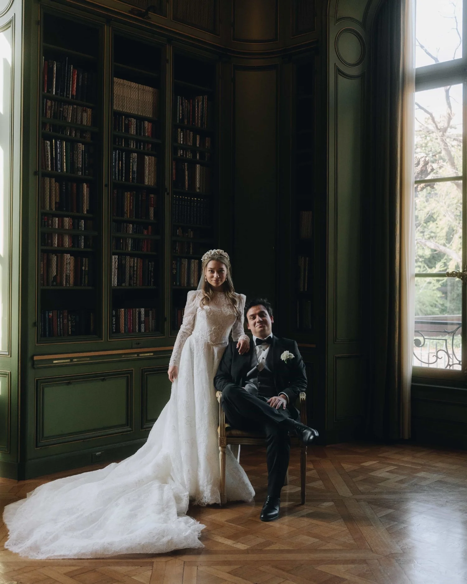 A bride standing beside the groom's chair in a green panelled library, her train laid out across the parquet