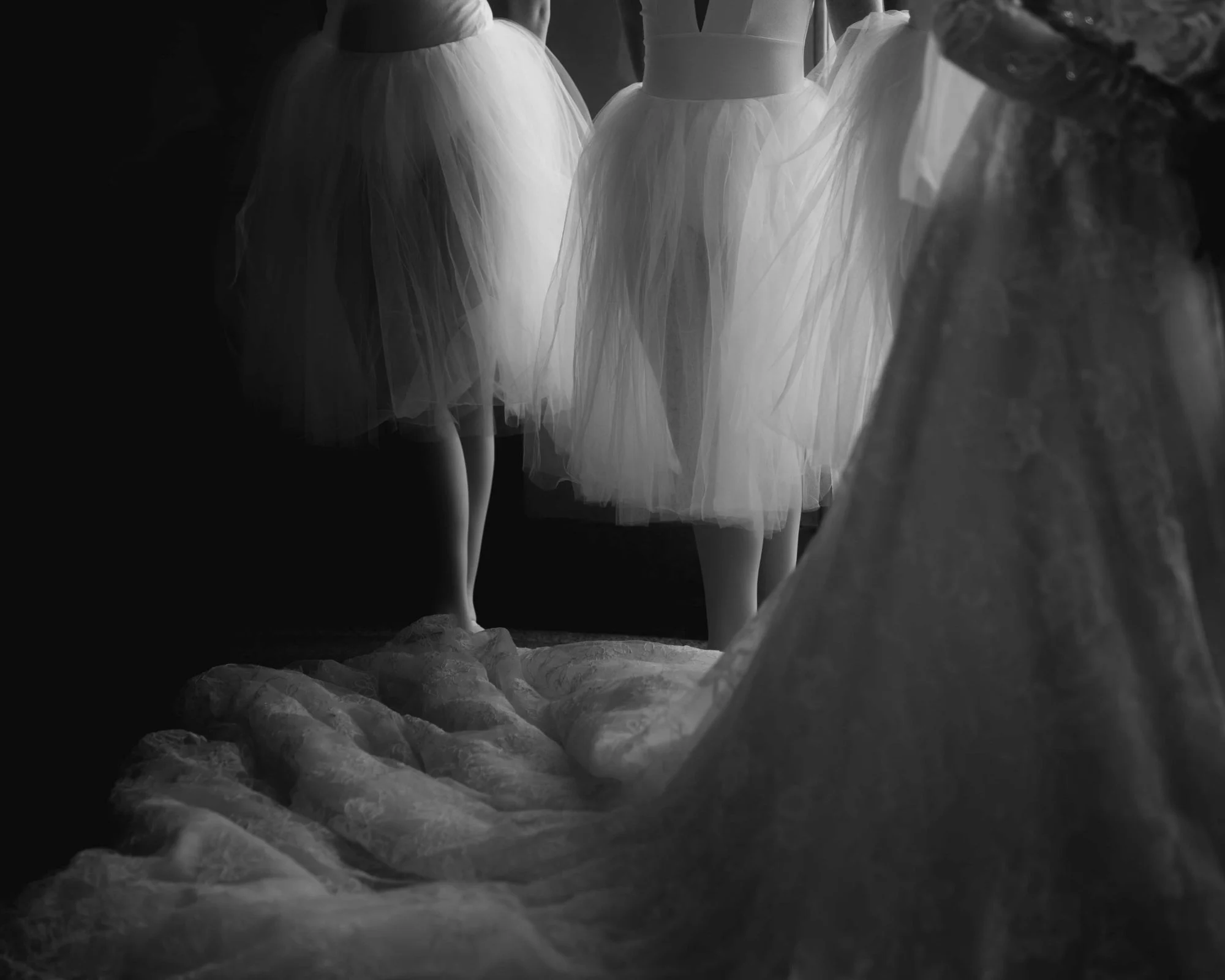 Black and white, dancers in tulle skirts standing barefoot at the edge of the bride's lace train where it pools across the floor