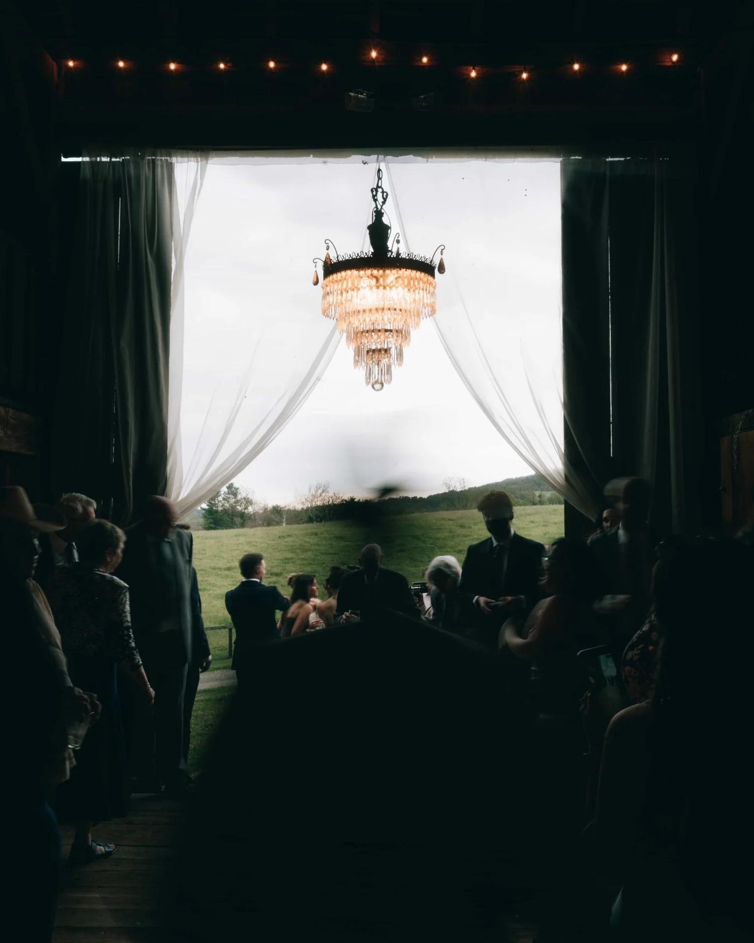 Looking down a dark aisle toward a bright draped opening with a chandelier hanging in it, the guests in silhouette